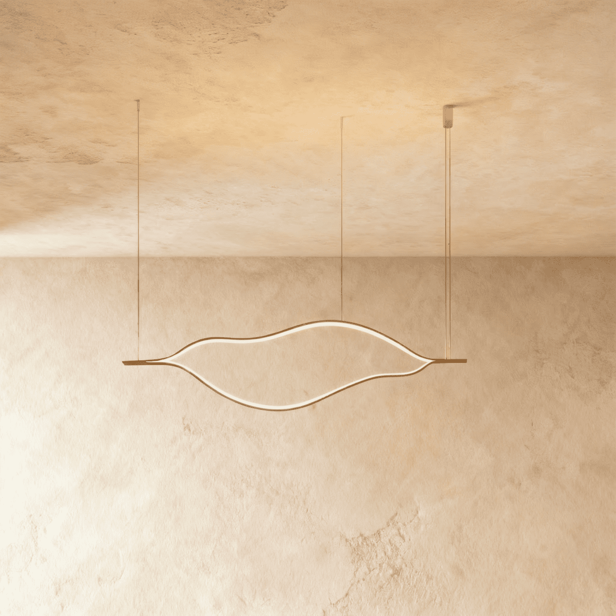Clérachic gold leaf-shaped linear chandelier switched on, suspended from slim cables in front of a softly textured limewashed wall.