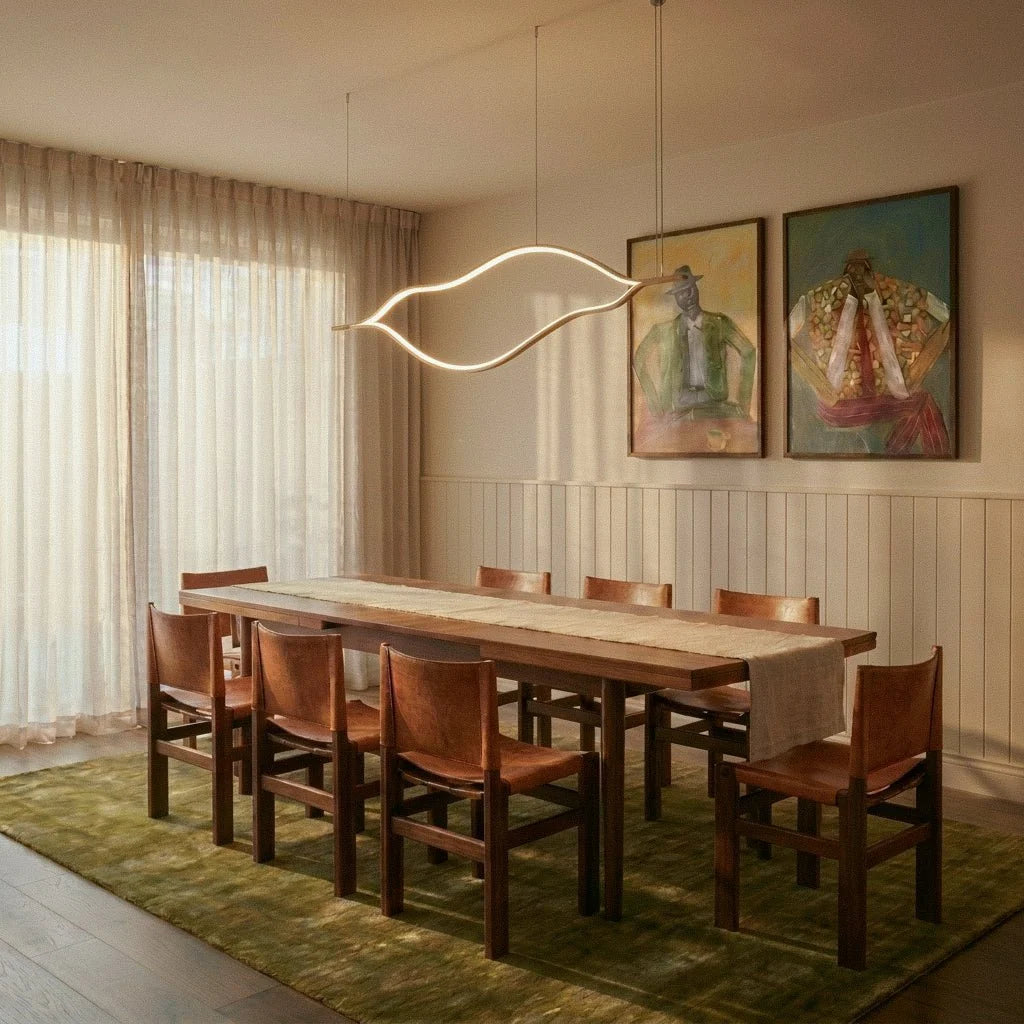 Clérachic gold wave-shaped linear chandelier above a long walnut dining table with leather chairs, beside sheer curtains and artwork.