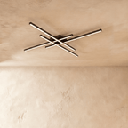 Clérachic black ceiling light of four crossed LED bars glowing warm white on a limewash ceiling, seen from below.
