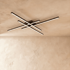 Clérachic black four-bar LED ceiling light lit warm, the crossed aluminium bars reaching wide across a limewash ceiling.