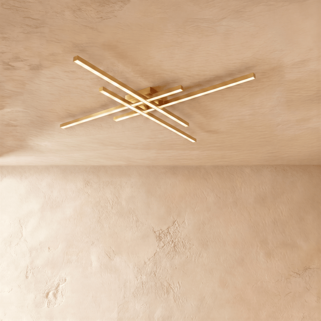 Clérachic gold four-bar LED ceiling light, slim brushed brass crossed arms glowing warm on a limewash ceiling.