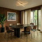 Clérachic gold crossed-bar LED ceiling light above a dark dining table with six chairs and walnut wall panelling.