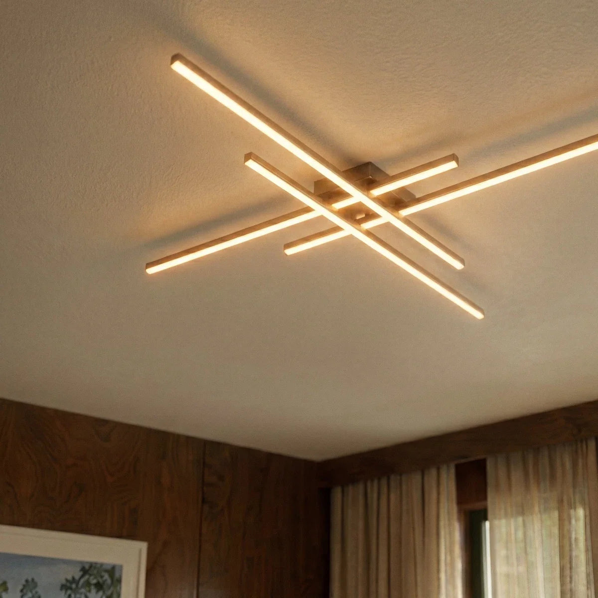 Clérachic gold four-bar LED ceiling light close up, glowing frosted bars on a ceiling beside walnut panelling and curtains.