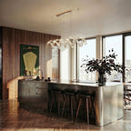 Clérachic spiral crystal chandelier on a brass bar over a steel kitchen island with stools, in a bright flat with tall windows.