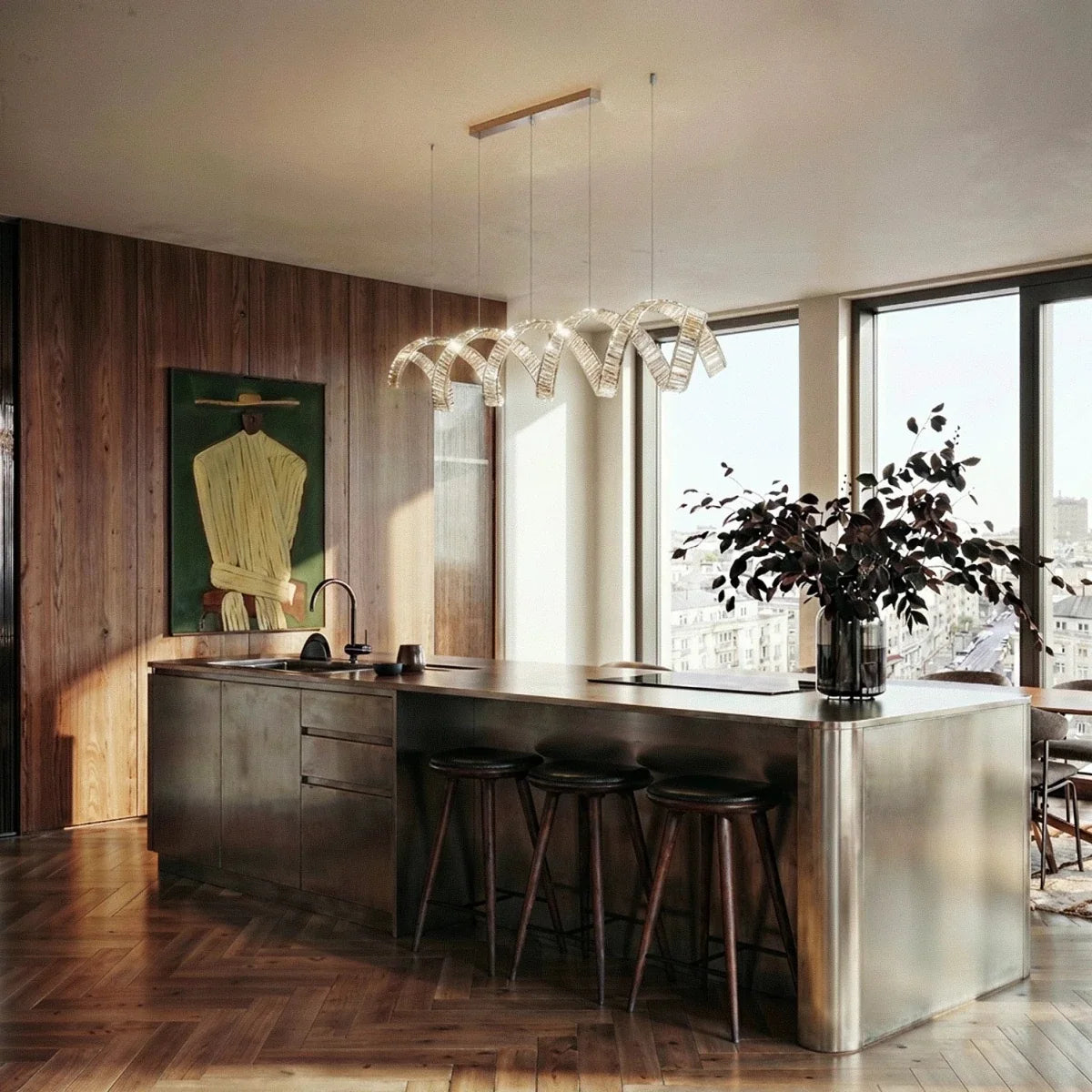 Clérachic spiral crystal chandelier on a brass bar over a steel kitchen island with stools, in a bright flat with tall windows.
