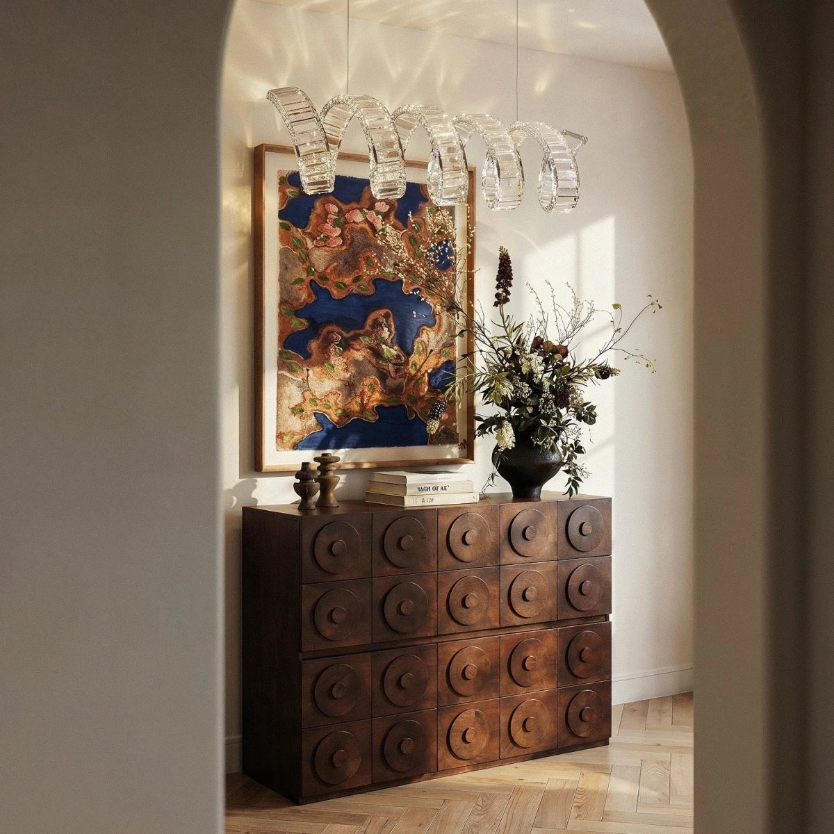 Clérachic clear-crystal spiral chandelier in an arched hallway above a carved dark wood chest and a framed artwork.
