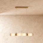 Clérachic linear pendant of four alabaster cylinders joined by brass bands, lit warm on two cords in a studio room.