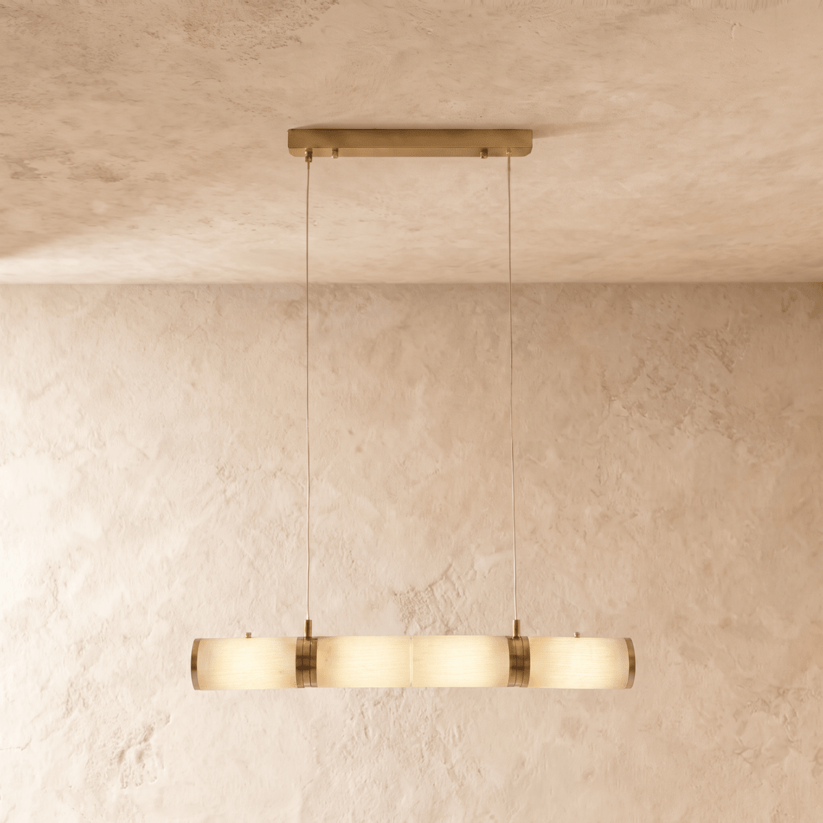 Clérachic linear pendant of four alabaster cylinders joined by brass bands, lit warm on two cords in a studio room.