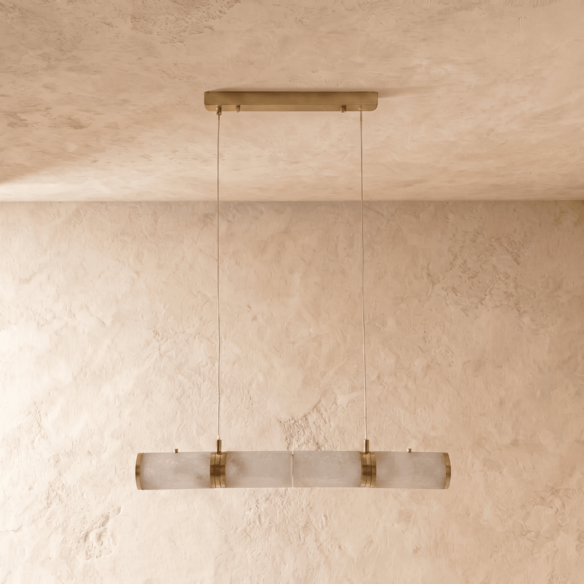 Clérachic linear pendant of four alabaster cylinders with brass bands, switched off, hung on two cords against limewash.
