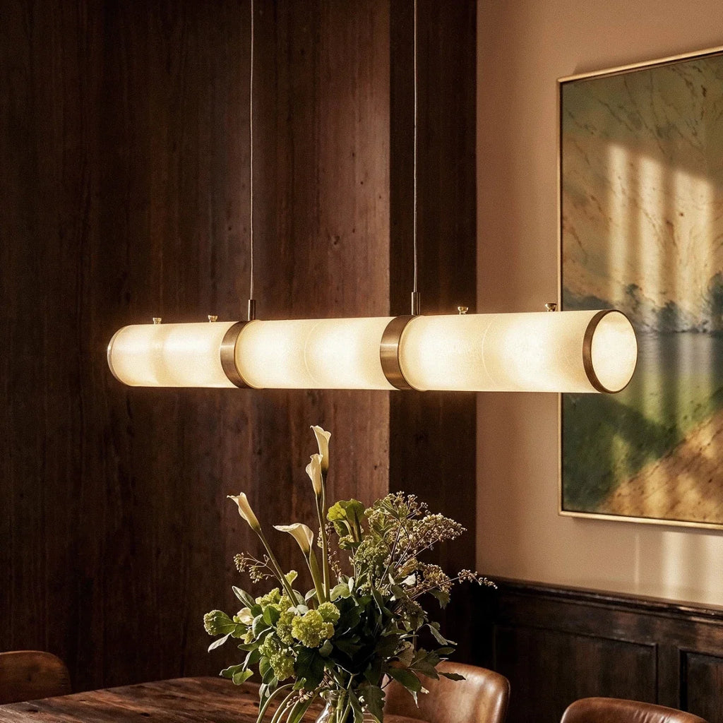 Close up of Clérachic alabaster cylinders with brass bands glowing warm above a vase of flowers in a dining room.