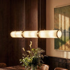 Close up of Clérachic alabaster cylinders with brass bands glowing warm above a vase of flowers in a dining room.