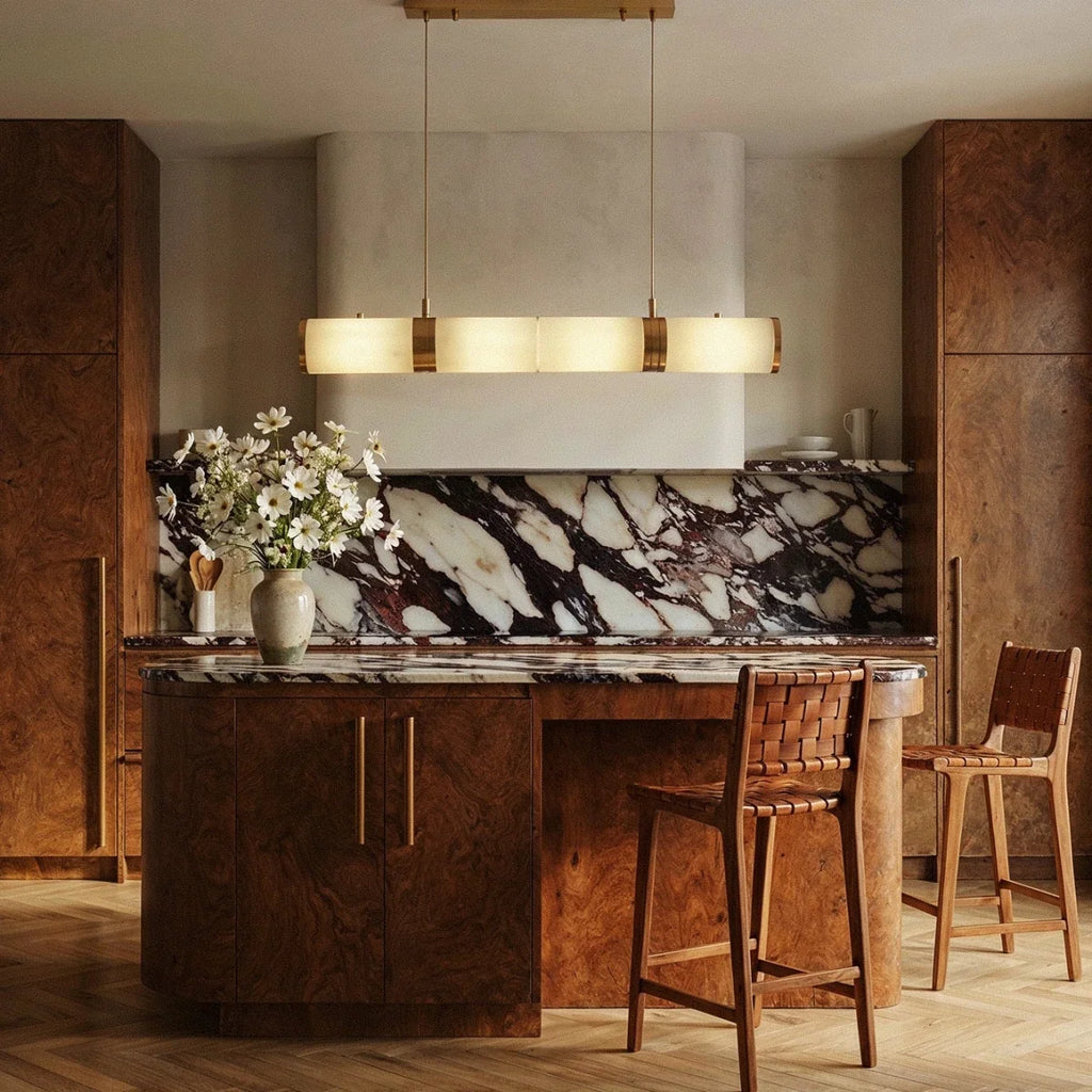 Clérachic linear pendant of glowing alabaster cylinders hung above a kitchen island with a marble splashback in a room.
