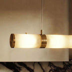 Close up of a Clérachic alabaster cylinder end cap, brass band and thin hanging cable, lit warm against a pale wall.