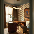 Clérachic alabaster linear pendant glowing above a curved wooden desk in a home office with patterned wallpaper.