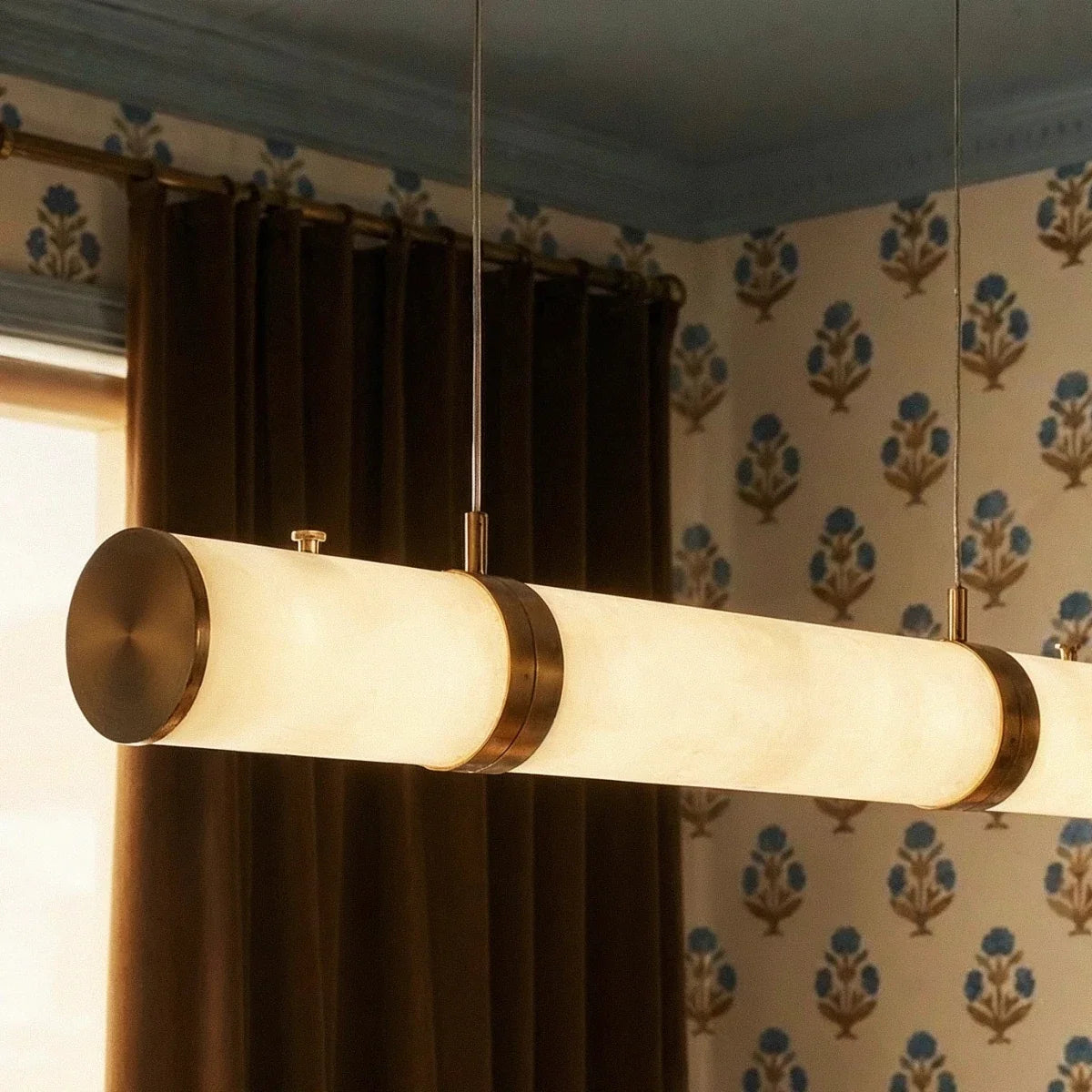 Close up of a Clérachic alabaster cylinder with a brass end cap and band, hung on two cables in a room with patterned wallpaper.