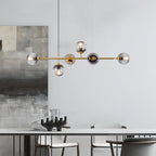 Lit Clérachic pendant of six glass orbs on a brass bar above a pale dining table, set against a soft grey wall with a framed abstract print.
