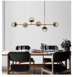 Above a walnut dining table with black leather chairs, this Clérachic linear pendant carries six smoky orbs on a brass rod.