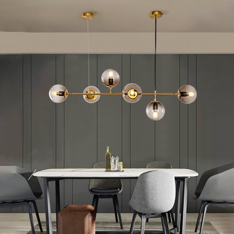 In a grey panelled dining room, a Clérachic brass pendant with six glowing glass orbs hangs over a white marble table.