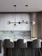 Six glass orbs on a slim black bar over a kitchen island with grey stools, in this Clérachic pendant on a marble-backed wall.