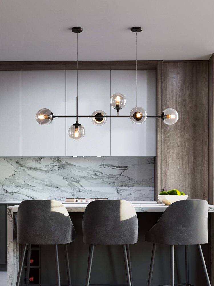 Six glass orbs on a slim black bar over a kitchen island with grey stools, in this Clérachic pendant on a marble-backed wall.