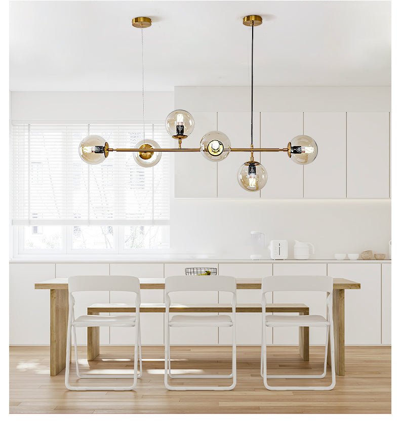 Over a pale wood dining table in a light kitchen, the brass rod and six clear amber orbs of this Clérachic pendant glow softly.