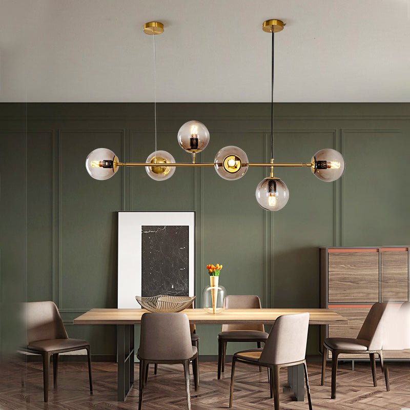 A Clérachic pendant with six lit glass orbs on a brass rod hangs above a wooden dining table against deep green wall panelling.