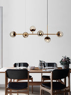 Six smoke-grey orbs along a brass bar, lit warm, over a set dining table with a jug and glasses, in this Clérachic pendant.