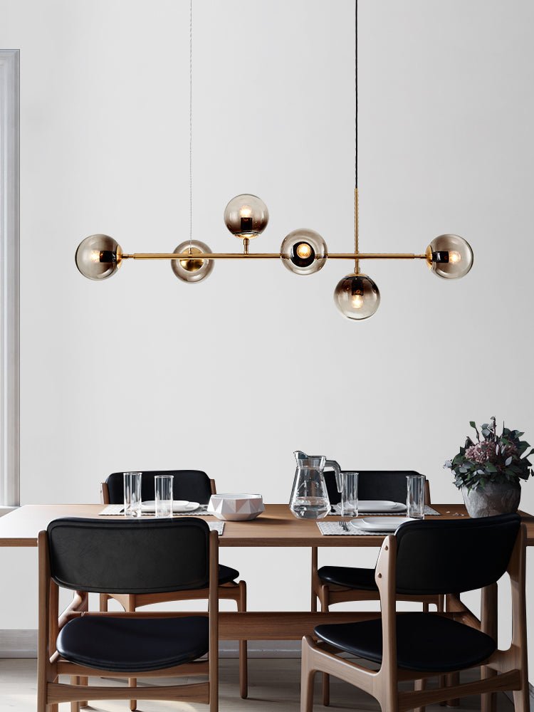 Six smoke-grey orbs along a brass bar, lit warm, over a set dining table with a jug and glasses, in this Clérachic pendant.