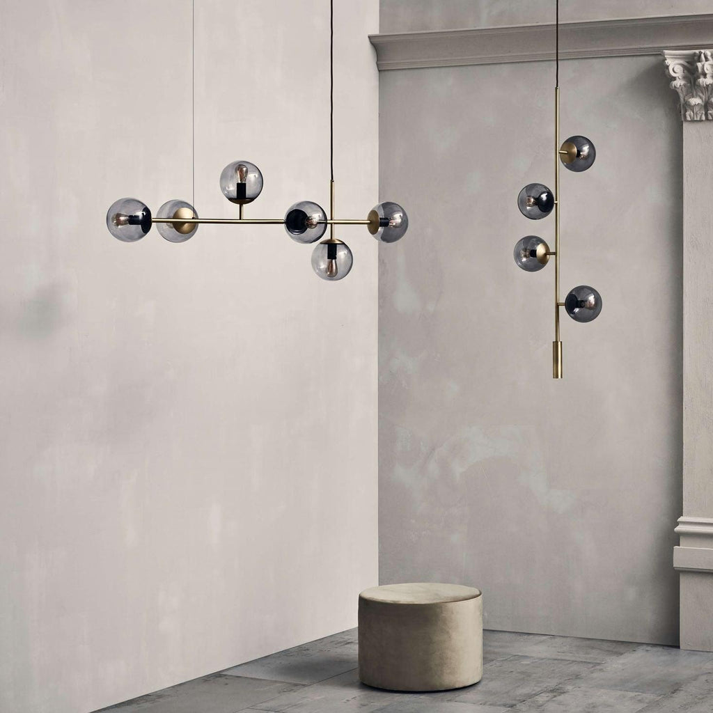 Smoked glass orbs on a brass bar hang in a plaster-walled room beside a round stool, a Clérachic pendant shown at room scale.
