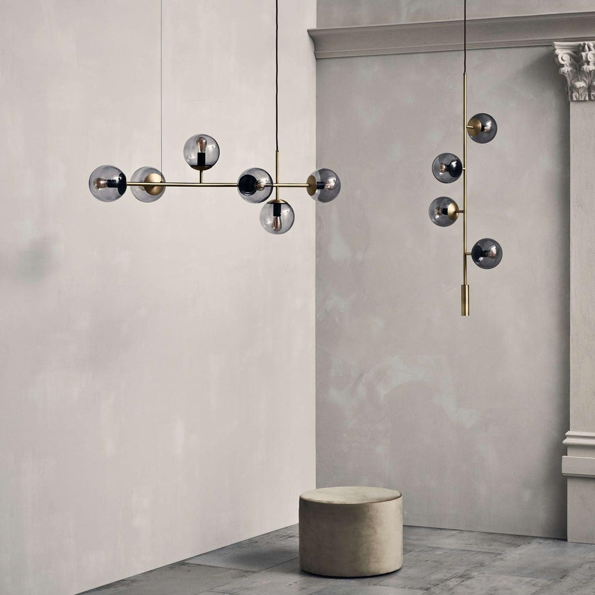 Smoked glass orbs on a brass bar hang in a plaster-walled room beside a round stool, a Clérachic pendant shown at room scale.