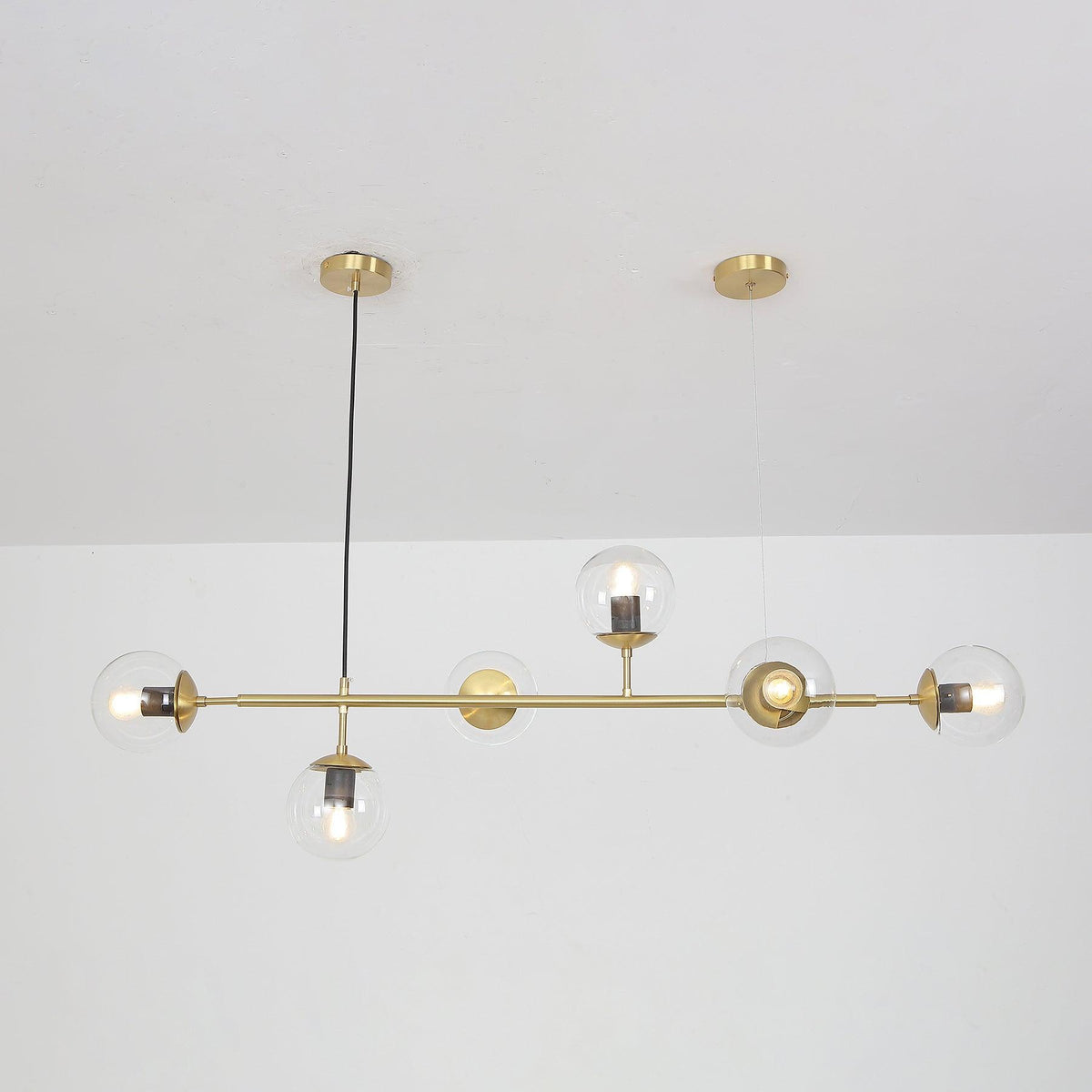 Clear glass orbs along a brass rod, hung from two round ceiling canopies against a white wall, in this Clérachic pendant.