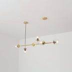 Lit from within, six clear glass orbs sit along the brass rod of this Clérachic pendant, shown against a white wall and ceiling.
