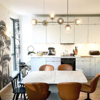 A Clérachic pendant with six smoked orbs hangs over a pale table in a kitchen with a botanical wall mural and tan leather chairs.