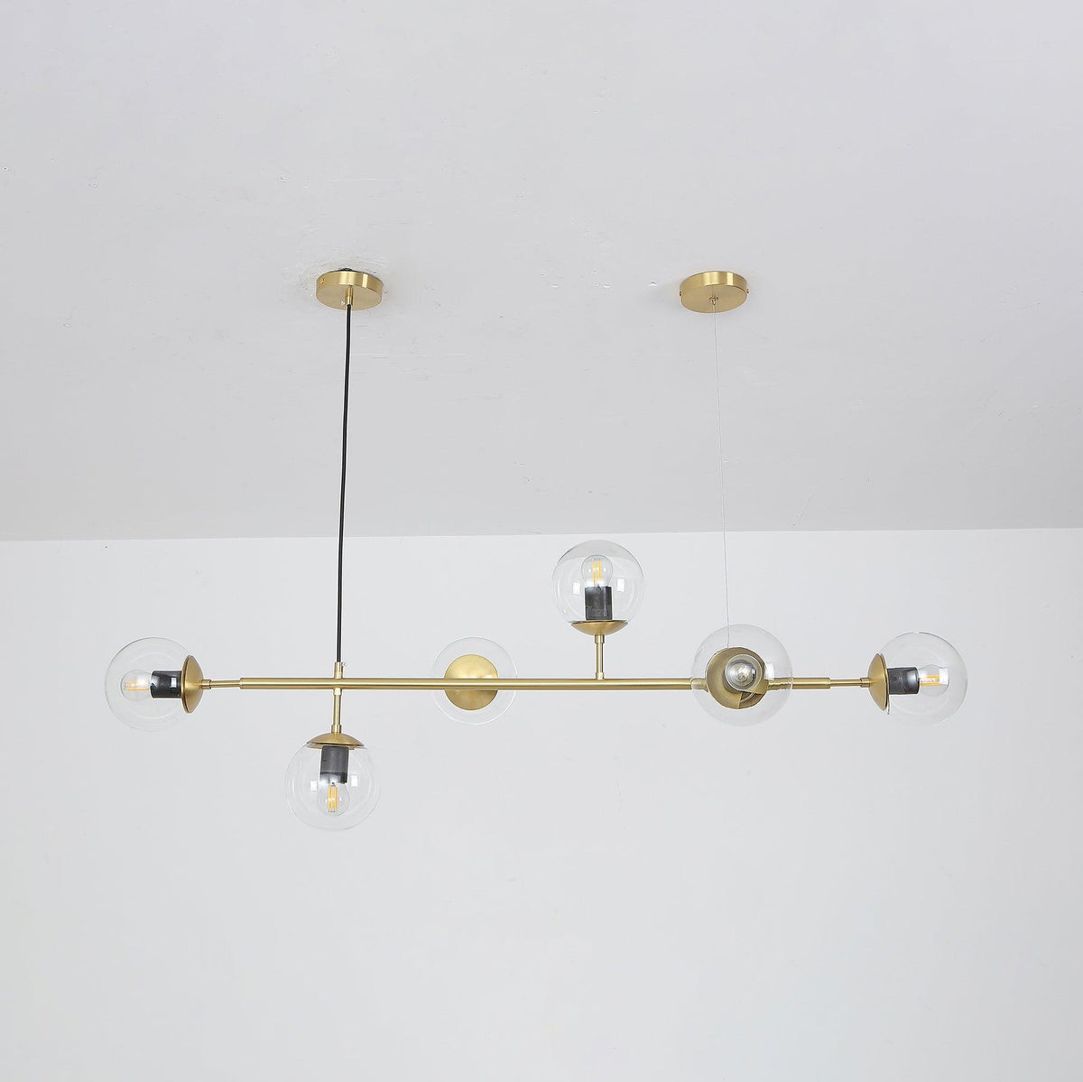 Clear orbs with their bulbs showing, strung along a brass rod from a single black cord, in a Clérachic pendant on a white wall.