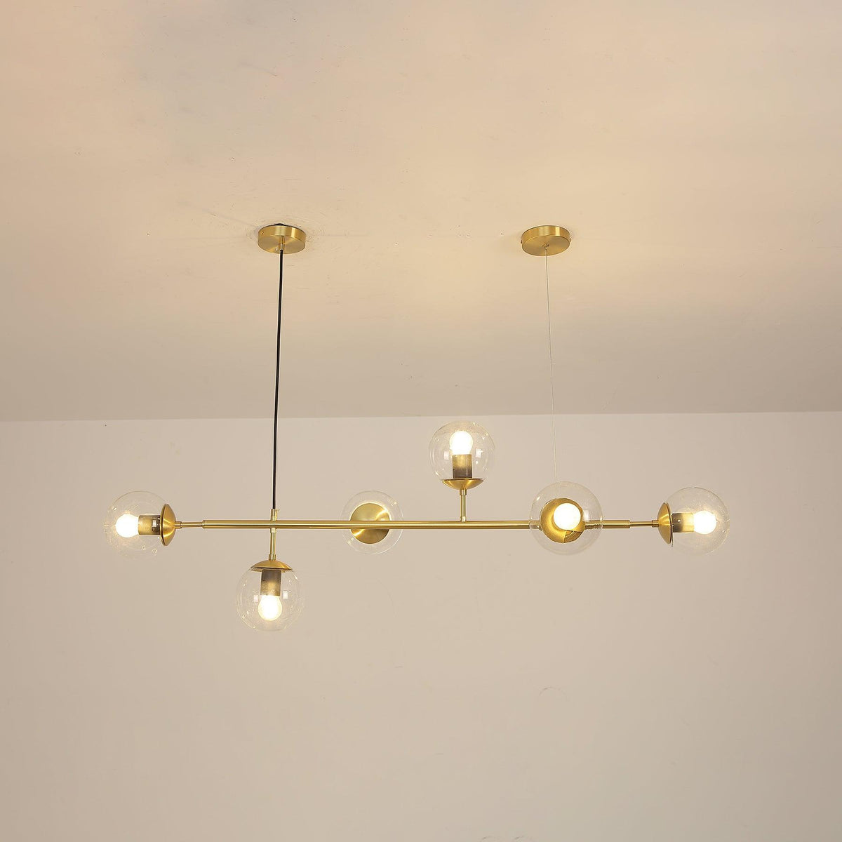 Switched on, the clear orbs of this Clérachic pendant cast a warm amber glow along the brass rod in a cream-toned room.