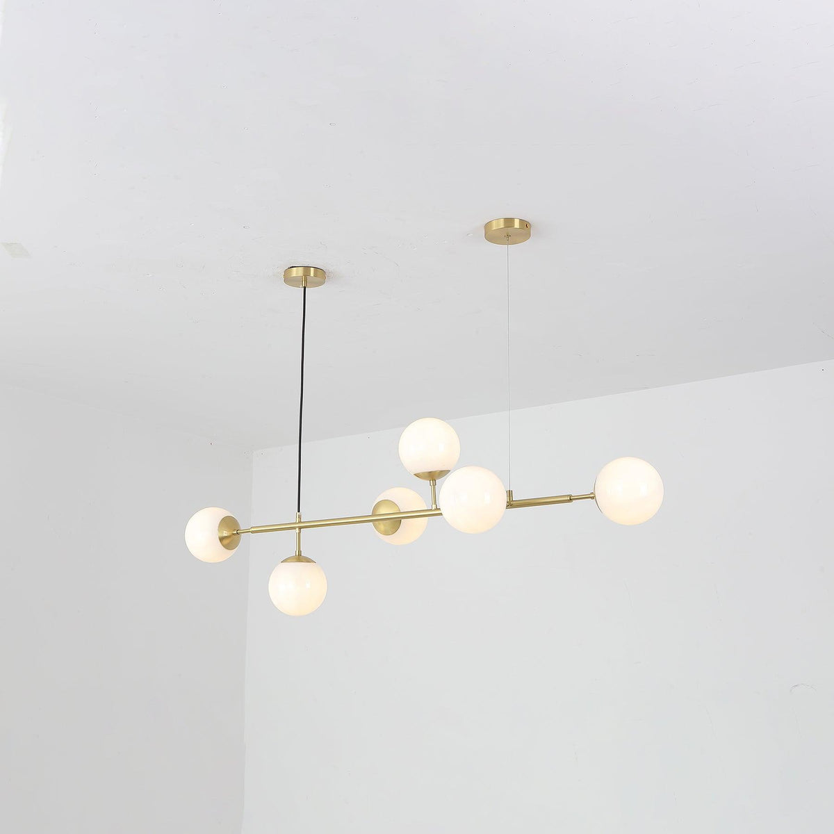 Six opal white orbs glow softly along the brass bar of this Clérachic pendant, seen from a low angle in a white room.