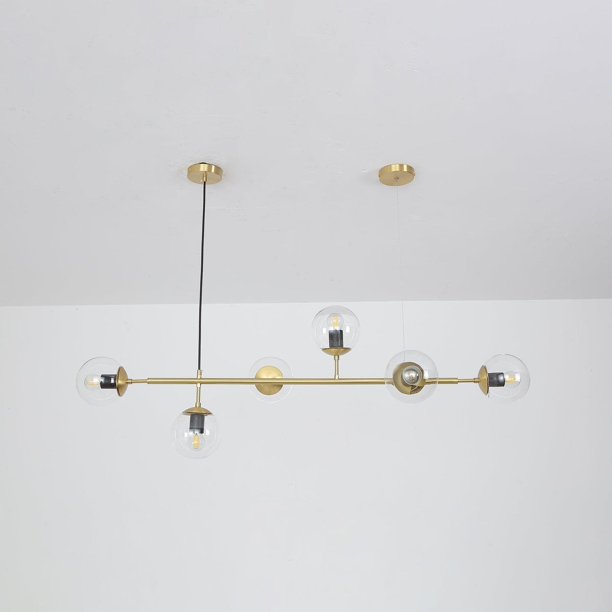 Straight-on view of this Clérachic pendant, its six clear orbs and bulbs hanging evenly from a brass rod below a white ceiling.