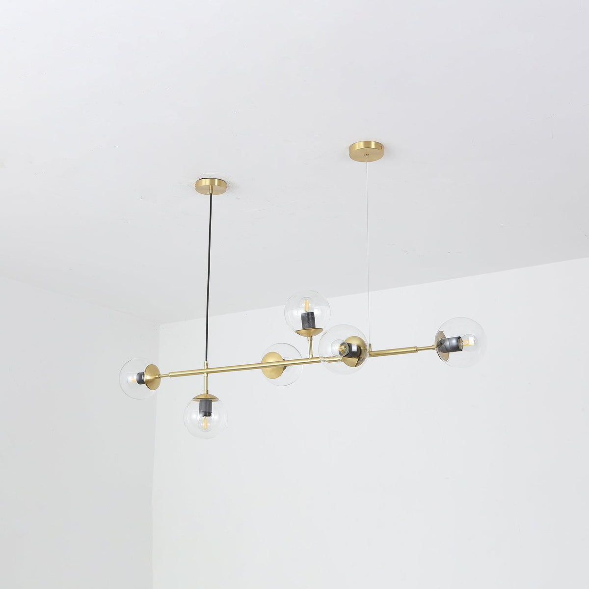 A Clérachic pendant on its brass rod, hung at a slight angle in a white corner, with clear glass orbs and visible bulbs.