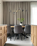 A Clérachic pendant of grey glass orbs on a thin brass bar hangs before floor-length grey curtains and a set oak table.