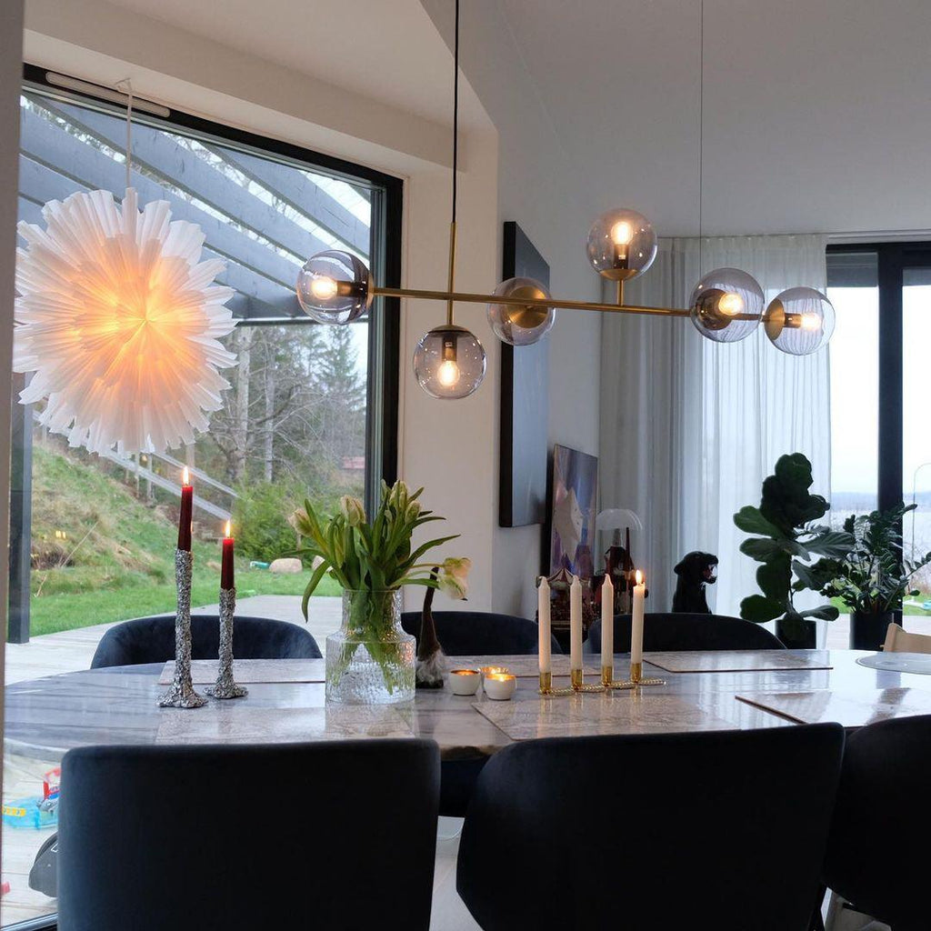Warm brass bar and smoky orbs of this Clérachic pendant glowing over a marble dining table, with candles and a paper star beside.