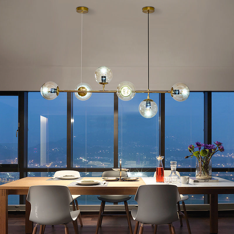 Clear amber orbs glow on a brass rod over a wooden dining table, with a night city view through floor-to-ceiling windows, from Clérachic.