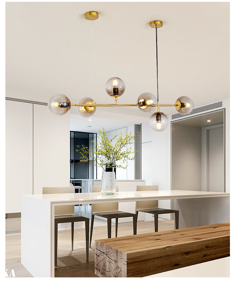 A Clérachic pendant with smoke-grey orbs on a brass rod hangs above a white island and wooden bench in a bright open kitchen.