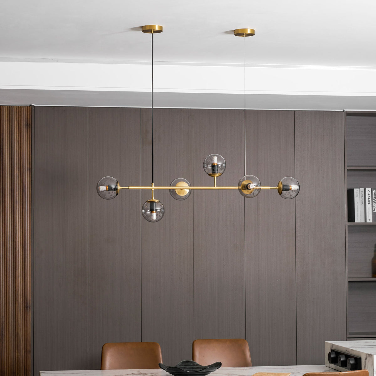 Lit amber glass orbs along a brass rod, over a dark wood panelled wall and a marble table, in this Clérachic pendant.