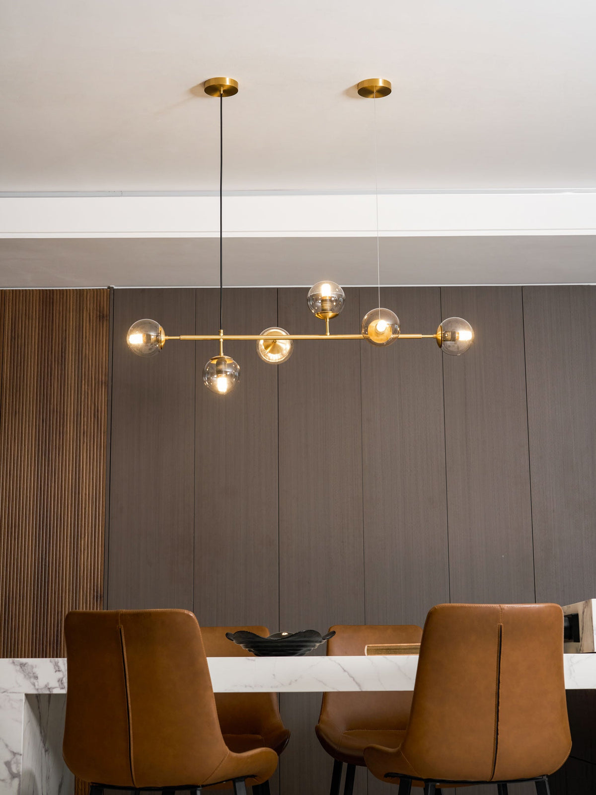 Six warm glowing orbs along a brass rod, hung before a dark panelled wall behind tan leather dining chairs, from Clérachic.