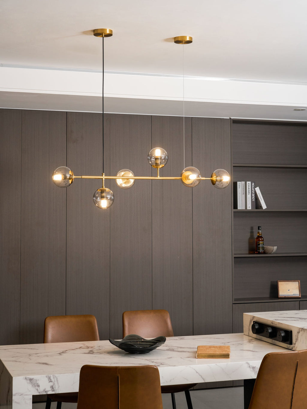 In a dining room with shelves of books and bottles, the brass rod and lit glass orbs of this Clérachic pendant glow warm.