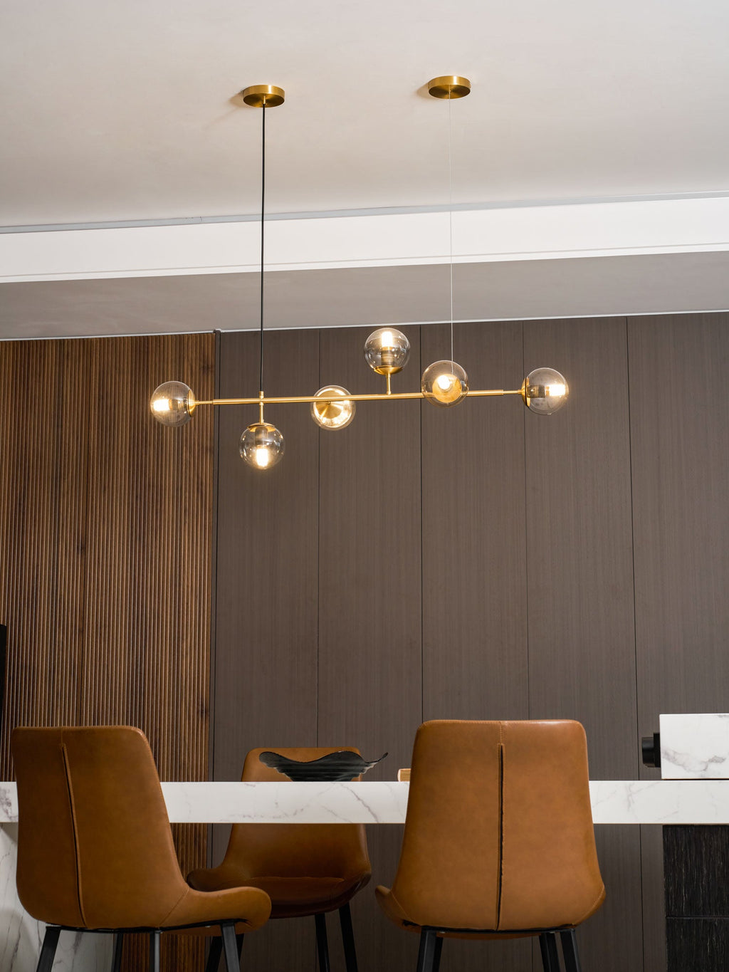 With a walnut panel at the left, this Clérachic pendant's lit orbs line up on a brass rod above a marble-topped table.