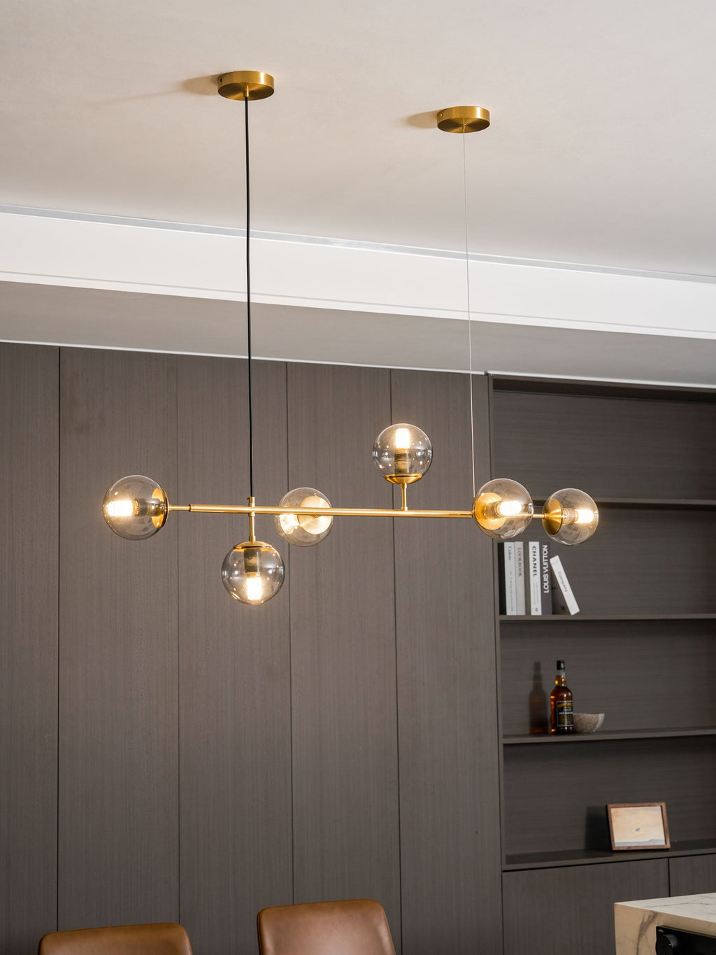A tall portrait view of this Clérachic pendant, its brass rod and lit orbs hanging over a panelled wall and bookshelf.