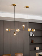 A tall portrait view of this Clérachic pendant, its brass rod and lit orbs hanging over a panelled wall and bookshelf.