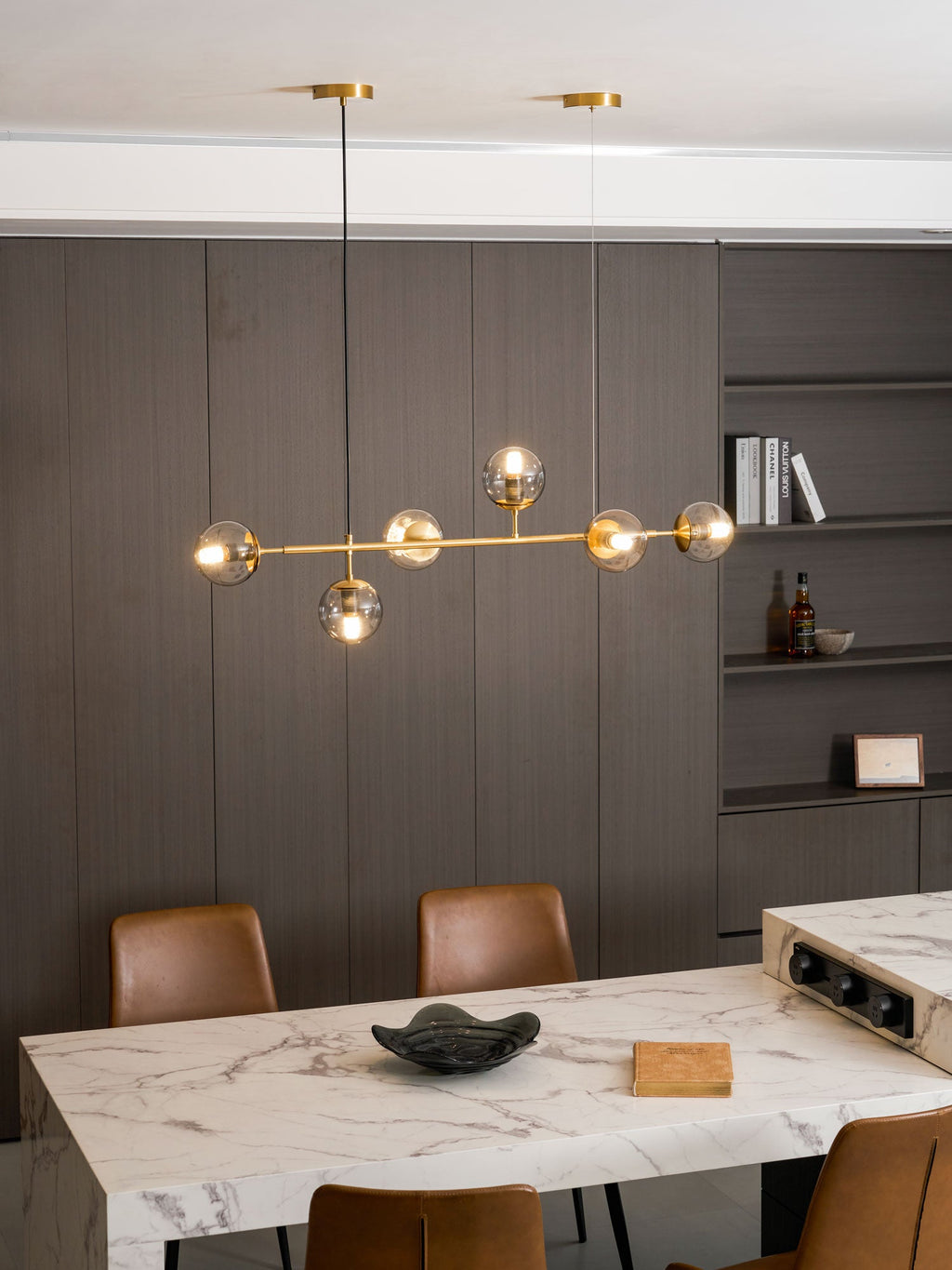 A full-length view of this Clérachic pendant, hung above a marble table with a green dish, with tan leather chairs below.