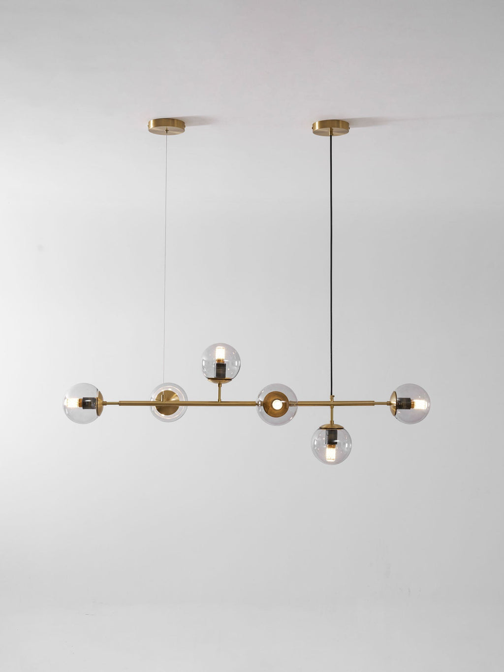 A smaller studio view of this Clérachic pendant on pale grey, six clear orbs lit along a brass rod from two brass canopies.